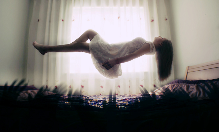 A woman, floating or levitating above a bed, dark lights reaching from the mattress while she floats in light.